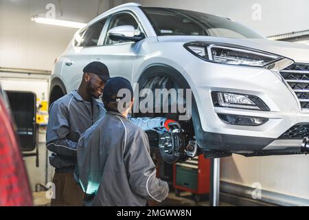 Zwei Mechaniker in der Nähe eines weißen Luxuswagens auf einer Hebebühne in einer Autowerkstatt, die Reifen repariert. Hochwertige Fotos Stockfoto