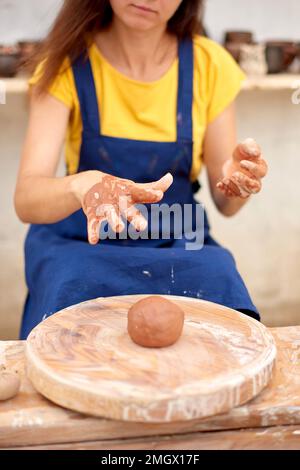 Handwerkerhände werfen ein Stück Lehm auf das Töpferrad. Gekürzte, nicht erkennbare Keramikfrau, die mit Töpferrad arbeitet. Stockfoto