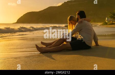 Ein wunderschönes junges Paar sitzt an einem tropischen Strand und genießt den Blick auf den türkisfarbenen Ozean während des Sonnenuntergangs Stockfoto