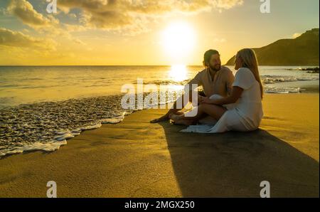 Ein wunderschönes junges Paar sitzt an einem tropischen Strand und genießt den Blick auf den türkisfarbenen Ozean während des Sonnenuntergangs Stockfoto