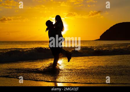 Silhouette eines Mannes, der eine Frau während der goldenen Stunde des Sonnenuntergangs an einem Inselstrand aufnimmt Stockfoto