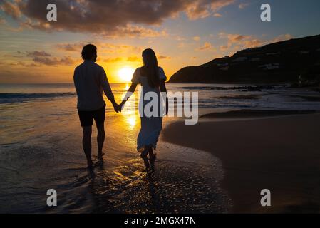 Ein junges, romantisches, verkleidetes Paar, das den Inselstrand hinuntergeht und den malerischen Sonnenuntergang ansieht Stockfoto