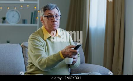 Weißer alter alter alter alter Mann in Brille entspannen Sie sich auf der Couch zu Hause, wechseln Sie den Sender mit Fernbedienung und sehen Sie sich ein Fernsehprogramm an, entspannen Sie sich beim Trinken Stockfoto