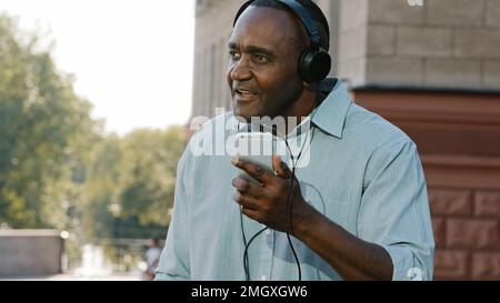 Emotional hochrangiger afroamerikanischer Mann, der in der Stadt draußen Musik über Kopfhörer hört und Musik hört. Glücklich erwachsen Stockfoto