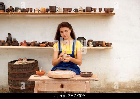 Professioneller Töpfer mit Tonklumpen in den Händen. Frauen arbeiten an Töpferrädern an weißen Wänden mit Regalen aus Tonschalen Stockfoto
