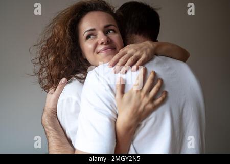 Junge, brünette, lockige Frau, die ihren Freund vor schlichtem Hintergrund umarmt und lächelt. Stockfoto