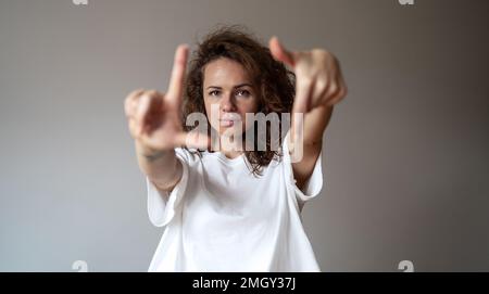 Junge braune, lockige Frau, die ein weißes T-Shirt trägt und mit den Fingern auf die Kamera schaut, vor einem einfarbigen grauen Hintergrund. Stockfoto