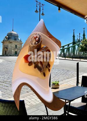 Die Budapester Brücke befindet sich in der Nähe eines Straßencafés in Ungarn. Sehen Sie den Wachturm und die Nahaufnahme der Dessertkreppiche in einem zusammengesetzten Bild, eine Spaziergang & Essensführung. Stockfoto