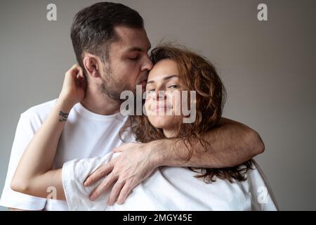 Porträt fröhliches junges Paar in weißer Kleidung, der eine Frau mit lockigem Haar umarmt und küsst, vor schlichtem Hintergrund. Stockfoto