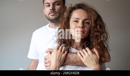 Porträt mit braunen Locken, Frau und bärtigem Mann, der hinter ihm steht und eine Frau mit der Hand umarmt, Paar, das weiße T-Shirts trägt und in die Kamera schaut. Stockfoto