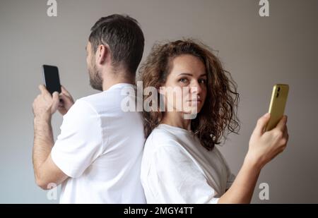 Frau und Mann, die weiße T-Shirts tragen, stehen Rücken an Rücken und benutzen ihre Handys getrennt, Frau schaut in die Kamera. Stockfoto