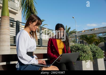 Zwei junge Geschäftsfrauen arbeiten im Freien, sitzen auf einer Bank und tippen mit ihrem Laptop. Digitales Nomadenkonzept Stockfoto