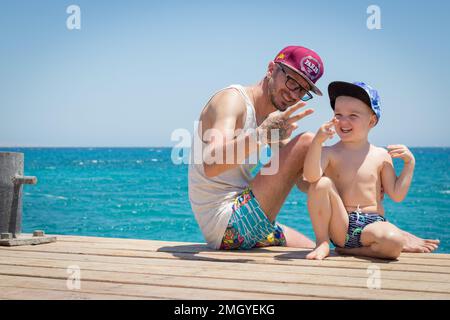 Portugal, Vilamoura, 02.01.2023. Vater und Sohn sitzen auf dem Pier und schauen auf den Ozean bei klarem, blauem Wetter. Das Konzept des Urlaubs Stockfoto