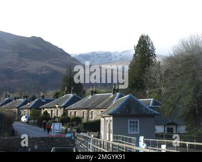 Reihen von Steinhäusern säumen die Pier Road im Naturschutzdorf Luss am Ufer des Loch Lomond mit einer spektakulären Bergkulisse. Stockfoto