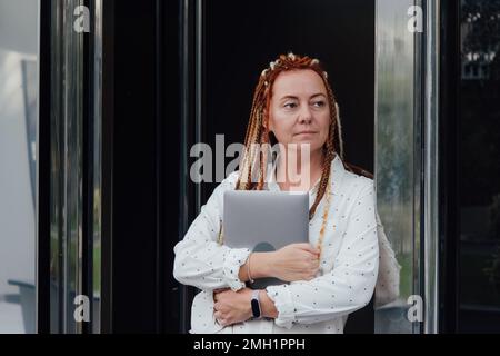 Eine Frau mit Dreadlocks und einem Laptop, die sich gegen die Haustür lehnt und auf ihre Uhr schaut Stockfoto