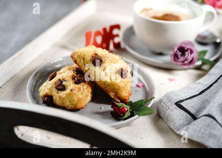 Frühstücksraum am Valentinstag - Scones Cappuccino Herz und Rosen auf einem Tablett, selektiver Fokus Stockfoto