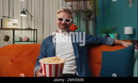 Ein Mann mittleren Alters mit Bart sitzt auf dem Sofa und isst Popcorn-Snacks und sieht sich zu Hause interessante Fernsehserien, Sportspiele, Filme und Online-Inhalte aus sozialen Medien an. Ein Typ genießt die Abendunterhaltung Stockfoto