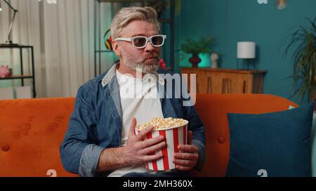 Aufgeregter älterer Mann sitzt auf der Couch und isst Popcorn-Snacks und sieht sich zu Hause interessante TV-Serien-Sportspiele, Filme und Online-Social-Media-Filme an. Ein älterer Mann genießt Abendunterhaltung Stockfoto