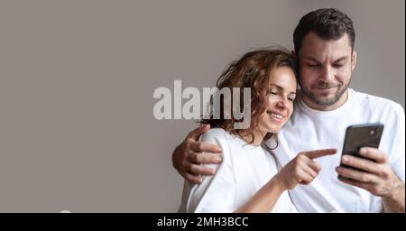 Ein junges Paar, das weiße T-Shirts trägt, eine lockige Frau und einen bärtigen Mann, zusammen mit einem Handy und einer lächelnden Frau, die auf den Bildschirm des Handys zeigt. Stockfoto