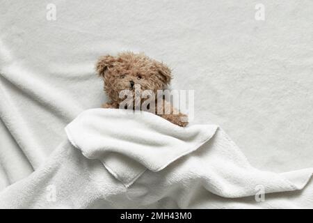 Ein brauner Teddybär liegt unter einer weißen Bettdecke auf einem Bett im Schlafzimmer, einem Kinderspielzeug Stockfoto