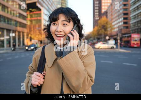 Portrait eines glücklichen koreanischen Mädchens, spricht auf dem Handy, sieht überrascht und glücklich aus, erhält positive tolle Nachrichten über Telefongespräche, steht fest Stockfoto