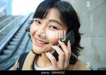 Nahaufnahme Porträt eines lächelnden brünetten Mädchens, koreanische Frau geht die Rolltreppe runter, telefoniert, spricht mit jemandem am Telefon Stockfoto