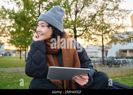 Lächelndes asiatisches Mädchen, die Schülerin sitzt allein im Park auf einer Bank, liest, nutzt die Social-Media-App auf einem digitalen Tablet, sieht sich Videos im Freien an, entspannt sich auf frischer Luft Stockfoto