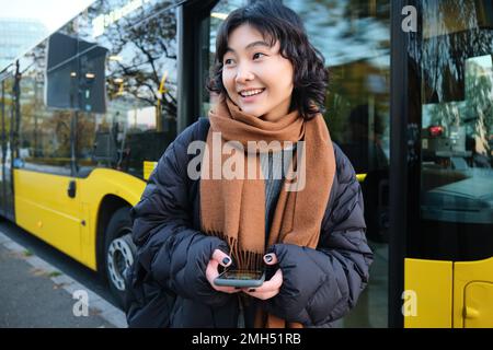 Junge, schöne Frau, die an der Bushaltestelle steht, SMS auf dem Smartphone schreibt, ihr Handy hält, ihren Terminplan überprüft, Fahrkarte online kauft, trägt Stockfoto