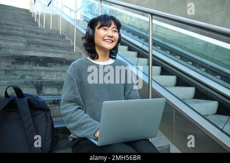 Female student works on project remotely, sits on stairs street, works on laptop, listens music in headphones and smiles Stockfoto