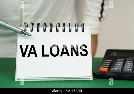 Geschäftskonzept. Auf einer grünen Oberfläche befindet sich ein Taschenrechner. Ein Mann zeigt mit einem Stift auf ein Notizbuch mit der Aufschrift „VA Loans“ Stockfoto