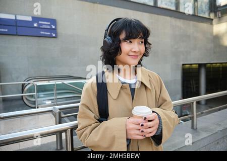 Porträt einer jungen Studentin, Mädchen mit Kopfhörern, trinkt Kaffee, steht auf der Straße mit Rucksack, pendelt zur Universität oder zur Uni, lächelt glücklich Stockfoto