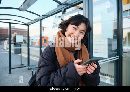 Süßes lächelndes asiatisches Mädchen, das an der Bushaltestelle steht, ein Smartphone in der Hand hält, eine Winterjacke und einen Schal trägt. Frau, die öffentlich zur Arbeit oder Universität pendelt Stockfoto