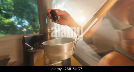 Das ätherische aromatische Öl von Hand aus einer Tropfflasche in Wasser für eine Kelle in einer hölzernen Fass-Sauna in norwegen. Dampfbad, Spa und Wellness Stockfoto