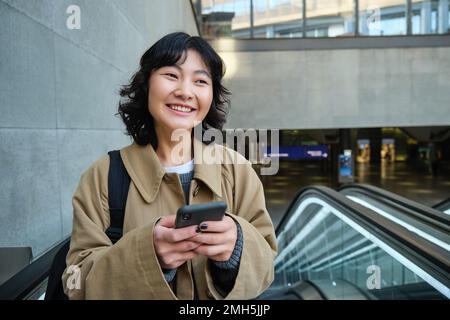 Porträt eines süßen koreanischen Mädchens im Trenchcoat, Rolltreppe hoch, hält Handy und lächelt zufrieden, pendelt Stockfoto