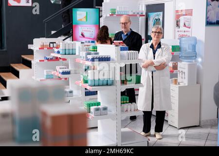 Porträt eines lächelnden älteren Apothekers, der in die Kamera schaut, während er in der Drogerie mit dem Arm überkreuzt steht und Kunden unterstützt. Kunden, die sich Apothekenregale ansehen, die Pillen und Vitamine kaufen Stockfoto