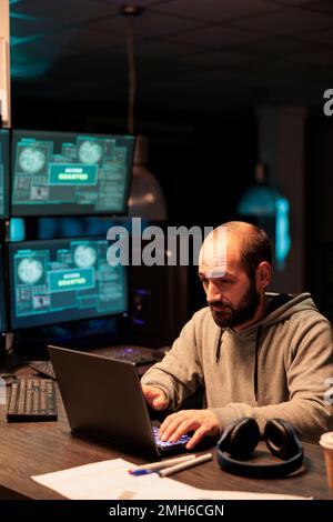 Gefährlicher Hacker, der im Büro in Datenserver eindringt und Computer und System mit Viren infiziert. Männlicher Dieb stiehlt Informationen von Software, arbeitet mit mehreren Monitoren. Stockfoto