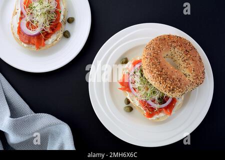 Frischer und gesunder geräucherter Lachs-Lachs-Lachs auf Sesamsamen-Mohn-Bagel mit Frischkäse, roten Zwiebeln, Sprossen und Kapern auf weißem Teller mit schwarzem Rücken Stockfoto