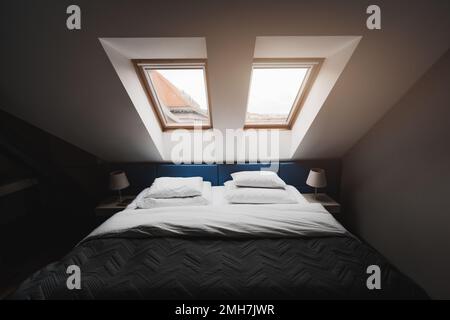 Ein Weitwinkel-Innenbild eines Schlafzimmers im letzten Stock mit zwei Fenstern; ein Hotelzimmer in einem Penthouse mit einem gemachten Bett und einer schwarzen Decke Stockfoto