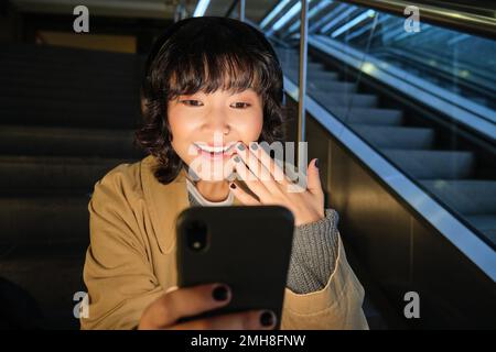 Das koreanische Mädchen sieht überrascht und glücklich auf ihrem Telefonbildschirm aus, lächelt erstaunt, liest gute Nachrichten, sitzt auf der Treppe neben der Rolltreppe im Einkaufszentrum, hört Musik Stockfoto