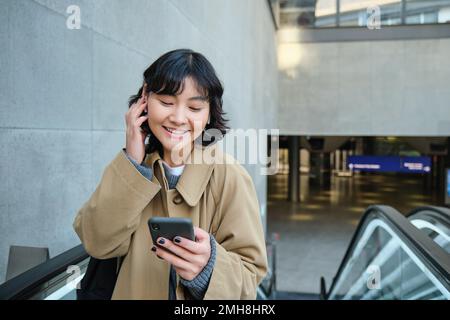 Junge brünette Frau pendelt zur Arbeit, geht irgendwo in die Stadt, steht auf einer Rolltreppe und benutzt ihr Handy, hält ihr Smartphone und lächelt, hört Musik Stockfoto