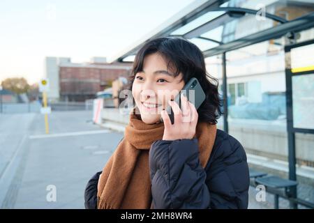 Lächelndes koreanisches Mädchen, das mit dem Handy spricht, an der Bushaltestelle steht, ein Smartphone benutzt, im Winter auf der Straße posiert, in einen Schal eingewickelt, eine schwarze Jacke trägt Stockfoto