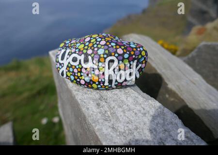 Freundlichkeit mit bemaltem „You Rock“-Schriftzug und bunten Punkten auf einer alten Steinmauer mit Klippen, die im Hintergrund nicht im Fokus sind Stockfoto