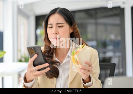 Geschäftige und hungrige asiatische Geschäftsfrau, die ein Sandwich isst, ihr schnelles Frühstück im Büro serviert und gleichzeitig auf die E-Mails von Geschäftskunden antwortet Stockfoto