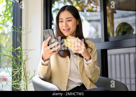 Eine wunderschöne und charmante asiatische Geschäftsfrau, die ihr Telefon benutzt, während sie ein Sandwich isst und im Café einen Brunch genießt. Stockfoto