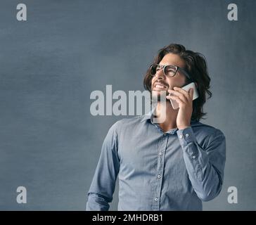 Ich rufe wegen Ihrer Anzeige an. Studioaufnahme eines jungen Mannes, der mit seinem Handy vor grauem Hintergrund spricht. Stockfoto