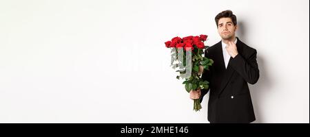 Bild von eleganten und frechen Mann in schwarzem Anzug, selbstbewusst und hält Strauß von roten Rosen, gehen auf ein romantisches Date, stehen gegen weiß Stockfoto