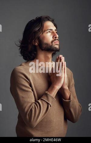 Ich könnte wirklich etwas Hilfe gebrauchen. Studioaufnahme eines gutaussehenden jungen Mannes, der vor grauem Hintergrund betet. Stockfoto