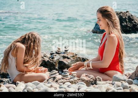 Eine Frau mit Tochter hat eine Biltsteinpyramide am Meer an einem sonnigen Tag auf blauem Meereshintergrund. Frohe Familienferien. Kieselstrand, ruhiges Meer. Konzept Stockfoto