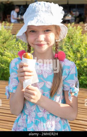 Hungriges Kind, das Straßenküche isst, sitzt auf einer Parkbank. Ein Mädchen, das gebratene Würstchen am Stiel isst. Glückliche Kinder, die Fast Food essen. Schnelle, kalorienreiche Straße Stockfoto