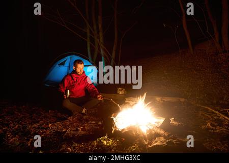 Hübscher junger Hipster, männlicher Tourist, der vor dem Lagerfeuer sitzt, mit gekreuzten Beinen und einer Tasse. Ein erwachsener, bärtiger Wanderer in der Nähe des Lagerfeuers bei Nacht Stockfoto